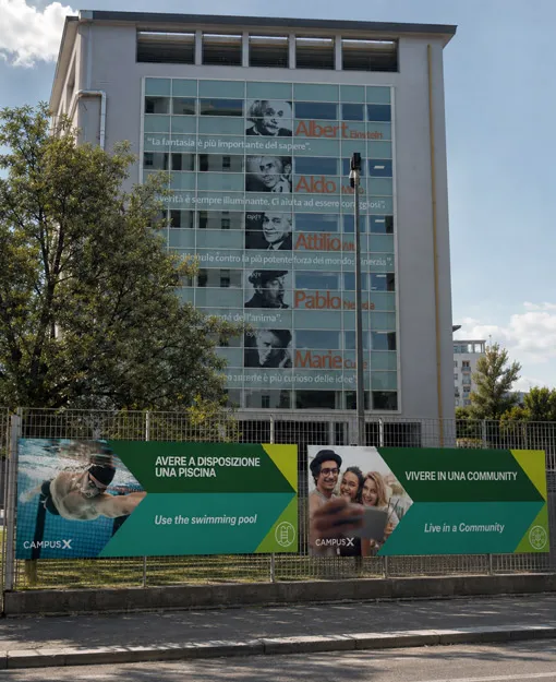 Two large-format advertising panels installed along the fence at the entrance to Campus X in Bari, set against a building with glass façades decorated with portraits and quotes from renowned figures such as Albert Einstein and Marie Curie: the first panel depicts an underwater swimmer with the tagline "Avere a disposizione una piscina / Use the swimming pool," the second shows a group of young people taking a selfie with the tagline "Vivere in una community / Live in a Community." Elena Lazzara designed the installation of the eleven advertising panels along the entrance path of Campus X in Bari, applying the brand's coordinated identity to the communication of the campus's spaces and services.
