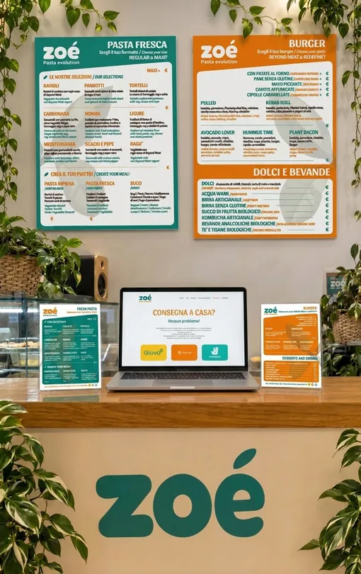 Comptoir de l'établissement ZOÉ avec logo vert, menus coordonnés vert et orange, supports d'information et ordinateur affichant le site web de la marque. Elena Lazzara développe un écosystème de branding pour un concept alimentaire plant-based, en intégrant identité visuelle, communication en point de vente et outils numériques au sein d'un système cohérent.un concepto alimentario plant-based, integrando identidad visual, comunicación en el punto de venta y herramientas digitales en un sistema coordinado.